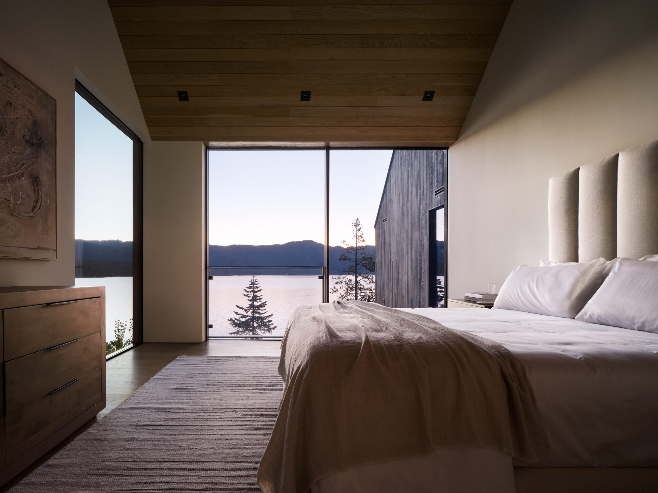 Cozy bedroom with a minimalist design featuring a wood-paneled ceiling, plush bed, and panoramah! minimalist sliding glass windows providing uninterrupted views of Lake Tahoe at sunset.